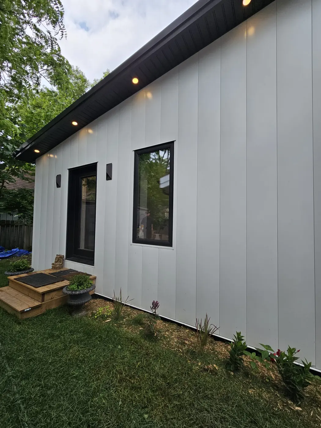Side elevation of the finished garden suite showing crisp vertical steel panel siding, black-framed windows, and new garden plantings