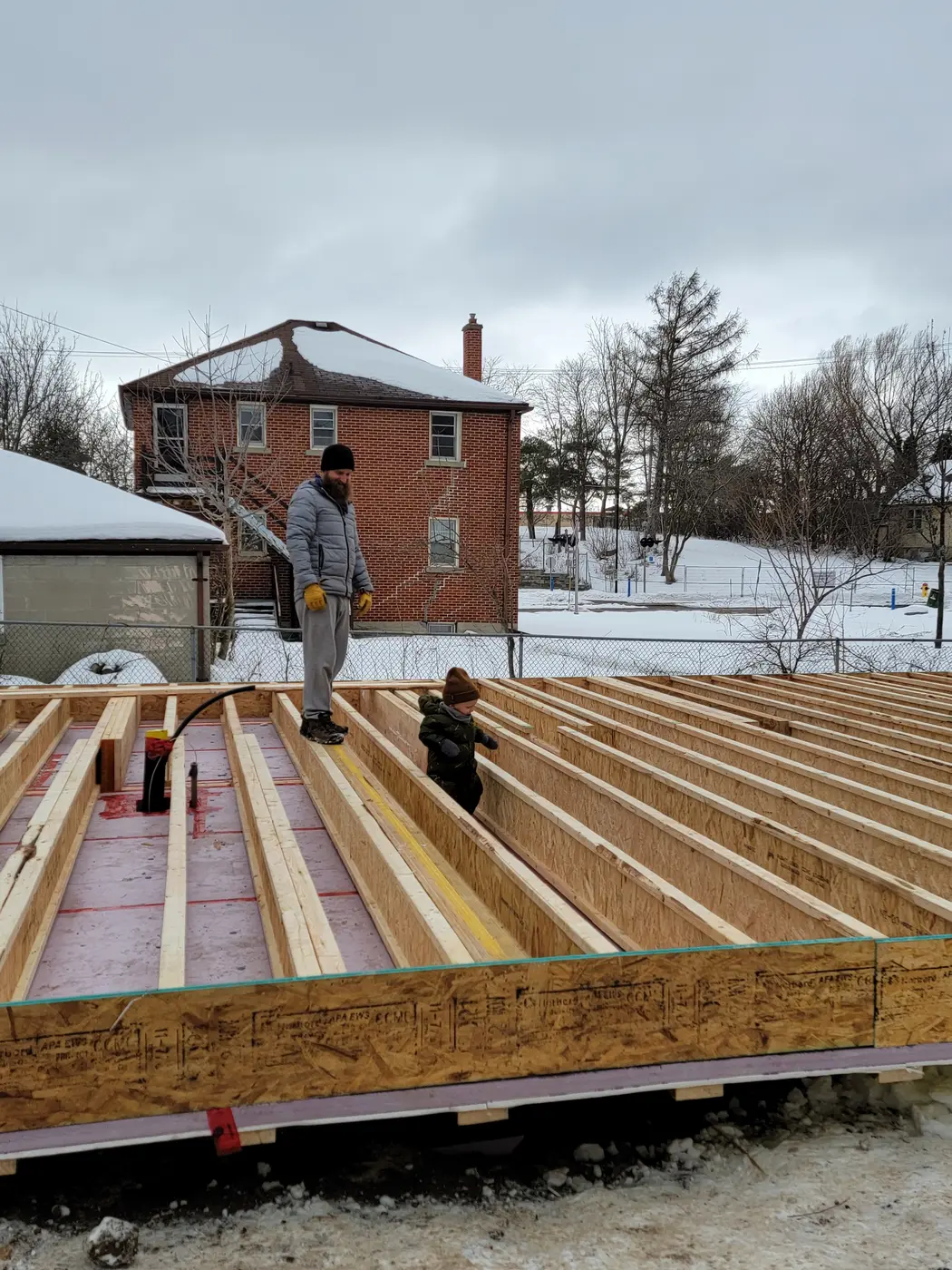 Two people standing on the open insulated floor joists of the garden suite in winter, with a brick neighbouring house behind
