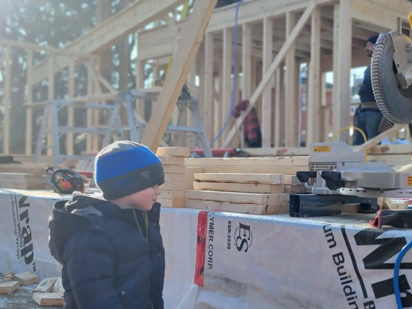 A young child in a winter hat watching a crew member work at a mitre saw on the construction site