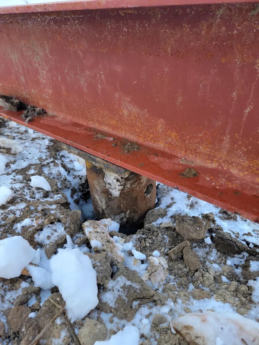 A steel helical pier rising from the ground, welded to a red structural steel beam that supports the floor system