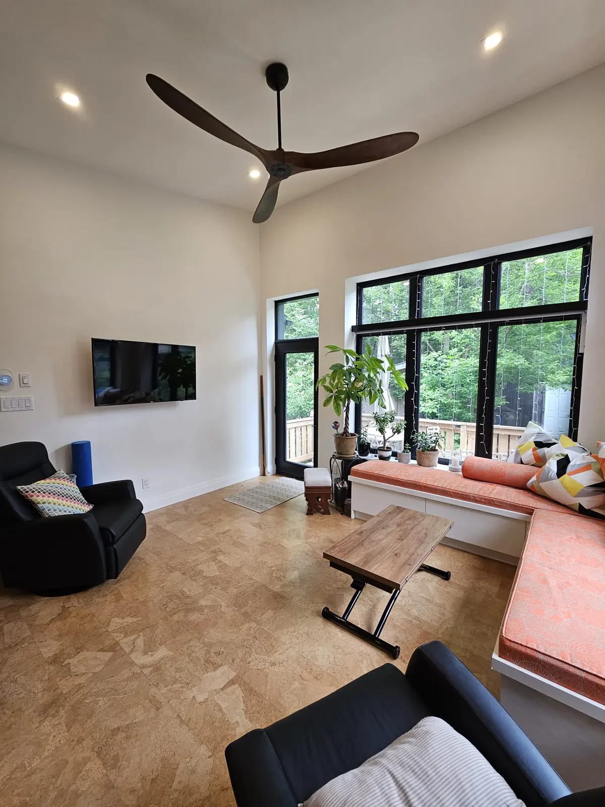 Open living area with cork flooring, a vaulted ceiling, a built-in window banquette, and large black-framed windows looking onto the garden